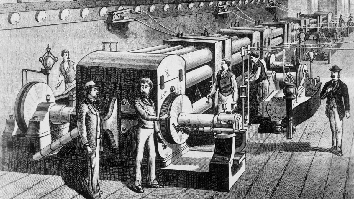 Black-and-white engraving of a long machine room with a row of large drum-shaped dynamos coupled to engines, men in suits and bowler hats standing beside them, and round lamps along the wall.