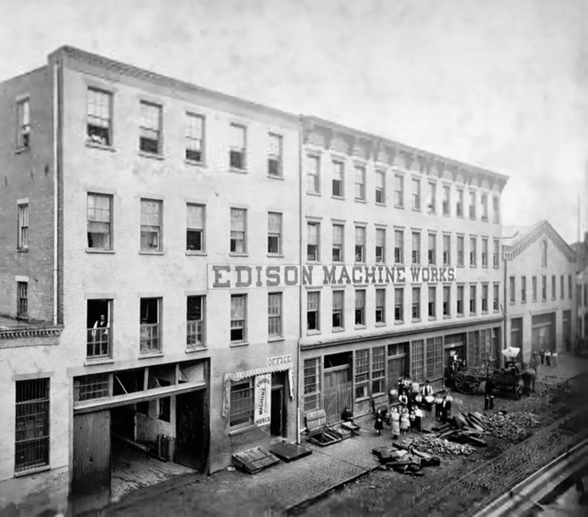 Black-and-white photograph of a long four-story brick factory on a cobbled street, with a painted sign reading Edison Machine Works across the front, a group of workers and a horse cart near the entrance.