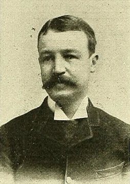 Halftone portrait photograph of a man with short dark hair and a thick mustache, wearing a dark jacket and a white collar.