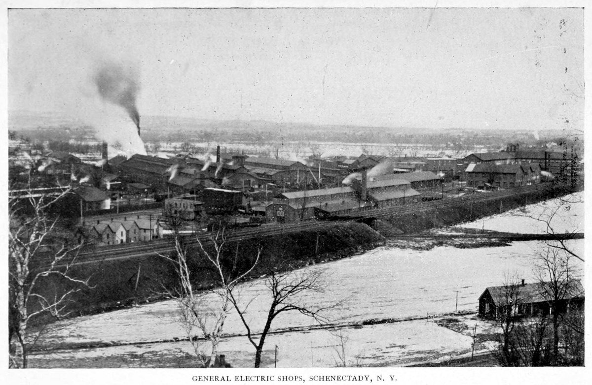Black-and-white photograph looking across a snowy river flat at a sprawling factory of long low sheds and smokestacks, with smoke rising and railway tracks in front.
