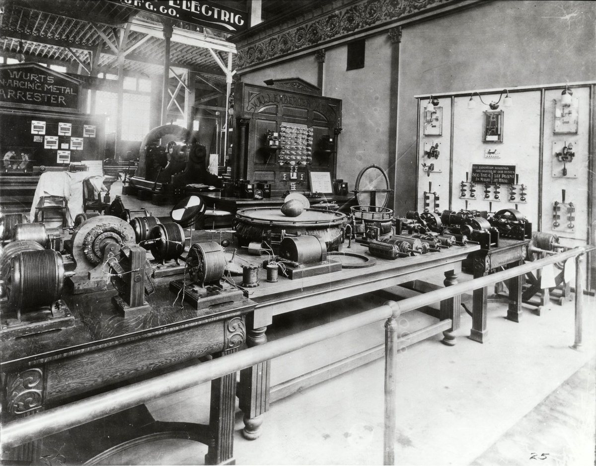 Black-and-white photograph of an exhibition stand with long wooden tables covered in electric motors and coils, a large ring with an egg-shaped object on it, and a switchboard panel.
