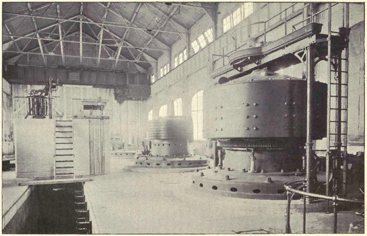Halftone photograph of a tall, bright machine hall with steel roof trusses and arched windows, where a huge drum-shaped generator stands on a round base and a second one stands half built behind it.