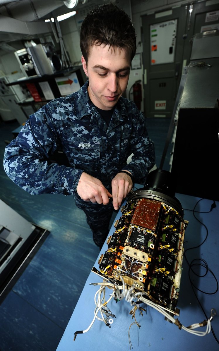 Color photograph of a technician in a blue camouflage uniform using small tools on an opened cylindrical aircraft generator full of wiring on a workbench.