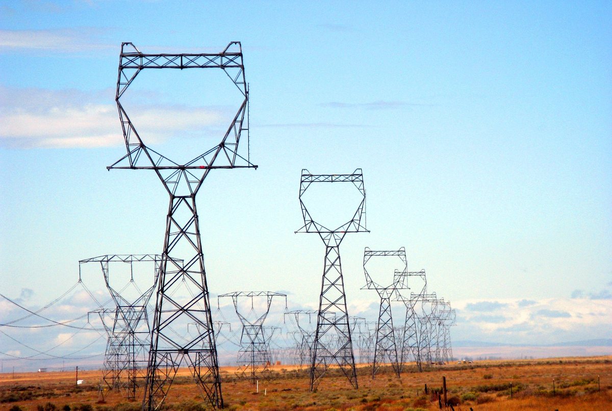 Color photograph of a row of tall steel lattice transmission towers carrying power lines across flat dry grassland under a blue sky.