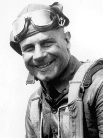 Photograph of Jimmy Doolittle smiling, in a leather flying helmet with the goggles pushed up, a leather jacket and a parachute harness
